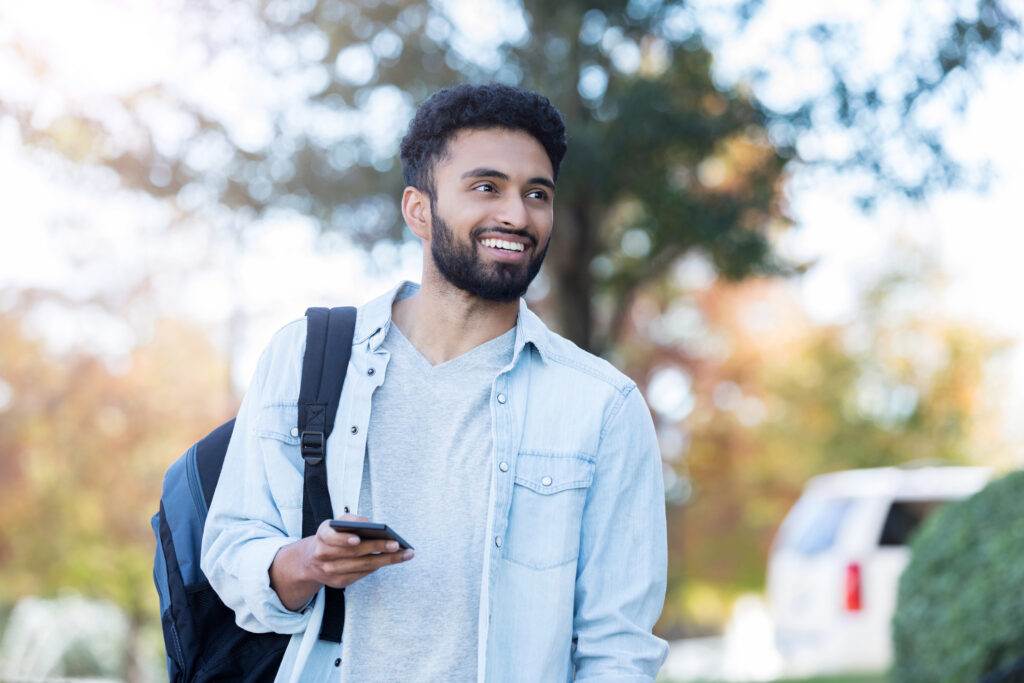 A smiling student walks outside on campus. They are holding a smartphone and carrying a backpack on one shoulder.
