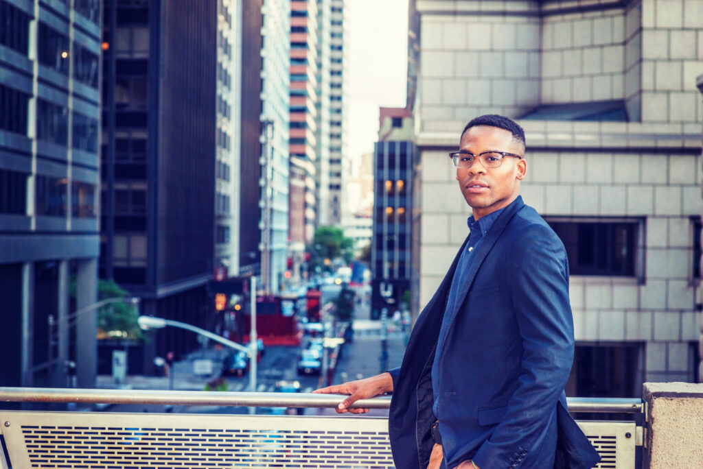 Wearing blue jacket, glasses, a young black man standing by railing on balcony, facing street, thinking, lost in thought. Instagram effect.