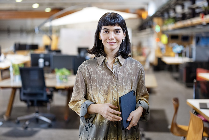 A woman in an office holding a tablet smiles at the camera