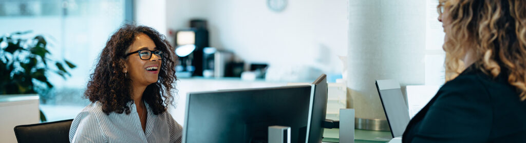 A woman at a computer smiles in conversation with another woman