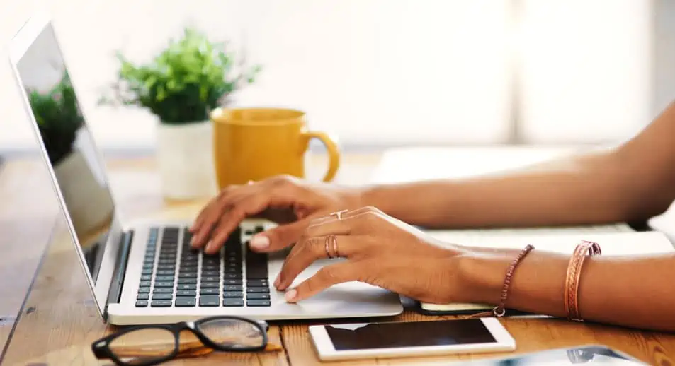 Hands typing on an Apple laptop. A pair of glasses and mobile device sit next to the laptop.